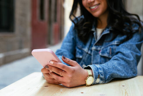 Crop unrecognizable young woman in casual clothes smiling and looking at her mobile phone screen while sitting at a table in a street cafe during a sunny day - ADSF45224