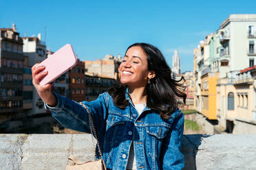 Ein lächelndes hispanisches Mädchen fängt einen Moment der Freude ein, während sie ein Selfie in ihrer Jeansjacke vor einer Stadtkulisse macht - ADSF45227