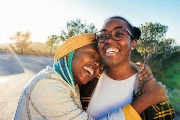 Laughing African American female with eyes closed embracing looking away ethnic friend in eyeglasses while standing together on road against green trees - ADSF45238