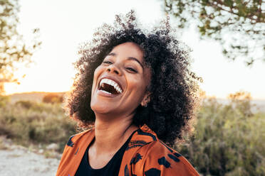 Portrait of young positive African American woman with curly hair laughing brightly standing against green grass - ADSF45240