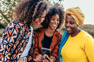 Young cheerful multiracial female friends in stylish clothes smiling and browsing smartphone while standing together - ADSF45242