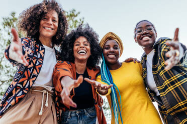 From below group of happy multiethnic female friends in stylish clothes smiling and looking at camera while standing under bright blue sky in park at daytime - ADSF45243