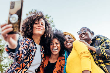 A diverse group of stylish female friends capture a joyful moment with a selfie under a bright blue sky in the park - ADSF45244