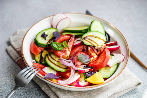 High angle of a tasty vegetarian salad with cucumber, lemon, onion and tomato with green leaves and dressing in a bowl placed on the table - ADSF45253