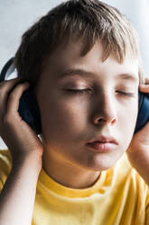 Portrait of serious eyes closed boy with brown hair wearing yellow t-shirt and touching headphones while listening to music against blurred background - ADSF45255