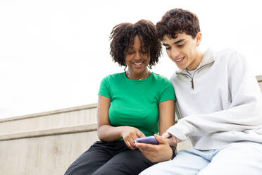 Smiling multiracial couple sitting on stairs against cloudless sky and looking at screen of mobile phone while enjoying interesting movie together in daylight - ADSF45266
