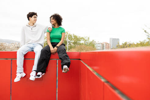 Confident multiracial couple sitting on red fence and looking at each other while male leaning on female's shoulder and resting in bright sunlight against cloudless sky - ADSF45267