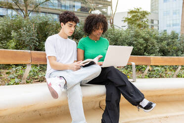 Concentrated young students sitting together on cement border with legs crossed in daylight while black female looking at screen of laptop and male reading book against green trees - ADSF45271