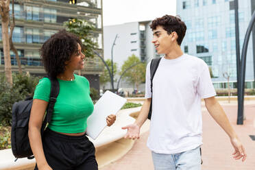 Positive multiracial male and female students with back packs standing in daylight on walkway and looking at each other while smiling and speaking in university campus with blurred buildings - ADSF45272