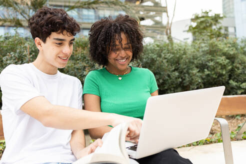Cheerful young students sitting together on cement border with legs crossed in daylight while looking and pointing at screen of laptop against green trees - ADSF45273