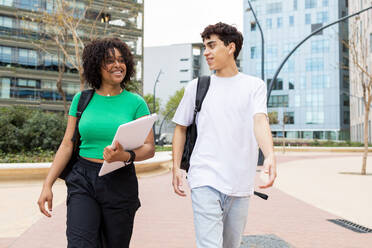 Positive männliche und weibliche Studenten mit Rucksäcken, die bei Tageslicht auf einem Gehweg spazieren gehen und sich lächelnd und sprechend auf einem Universitätsgelände mit unscharfen Gebäuden ansehen - ADSF45274