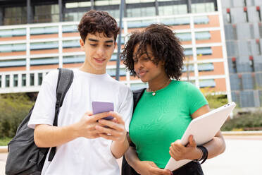 Positive young multiracial male and female students with backpacks looking at screen of mobile phone while standing together in daylight near modern building on college campus - ADSF45275
