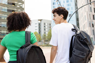 Back view of smiling young male and anonymous female with curly hair looking at each other while walking with backpacks on college campus road with buildings in daylight - ADSF45276