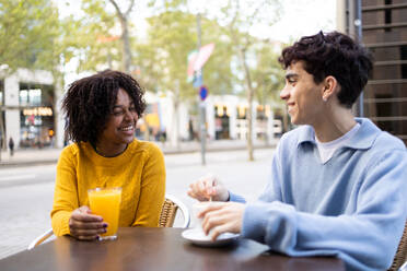 Smiling young multiracial student friends sitting at table with orange juice glass and coffee cup in cafe while looking at each other and speaking against blurred campus - ADSF45278