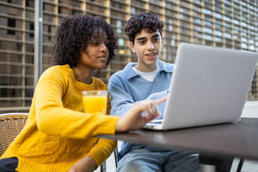 Multiracial young student friends sitting at table with orange juice glass and looking at screen of laptop in daylight while black female pointing finger on monitor and discussing with male - ADSF45279