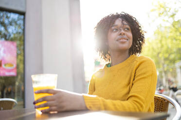 Positive young African American female with curly hair smiling and looking away while sitting at table - ADSF45282