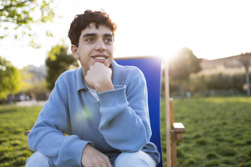 Cheerful young male in casual clothes sitting on deckchair with hand at chin looking away while relaxing in green park on sunny day - ADSF45291