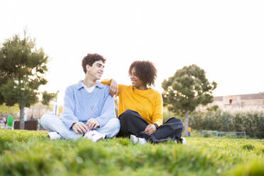 Smiling multiracial young female leaning arm on male's shoulder while sitting on green grass in park and looking at each other in daylight - ADSF45293