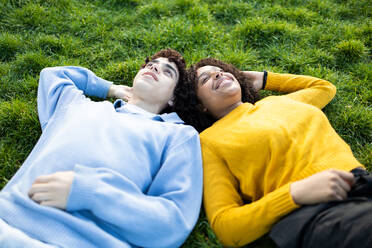 From above smiling multiracial young male and female friends lying on green grass with hand behind head in park and looking up in daylight - ADSF45294