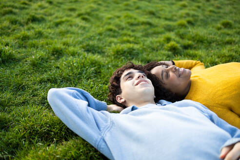 From above smiling multiracial young male and female friends lying on green grass with hand behind head in park and looking up in daylight - ADSF45295
