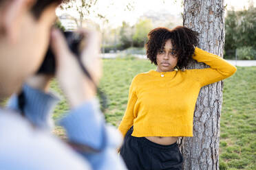 Blurred crop young male photographer shooting pictures of African American female standing near tree in lawn with hand behind head while looking at camera and posing in daylight - ADSF45298