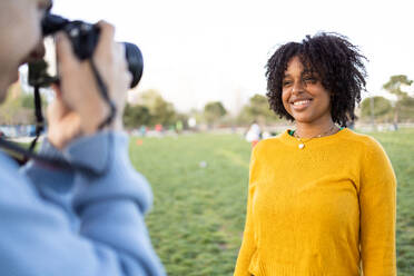 Blurred crop young male photographer shooting pictures of African American happy female standing against blurred background while looking at camera and posing in daylight - ADSF45299