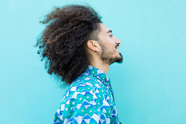 Side view of bearded African American male model with curly hair and earrings standing against blue background while looking away - ADSF45307