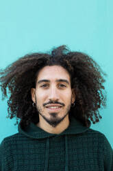 Portrait of positive African American male with curly hair in warm sweater smiling and looking at camera against blue wall background - ADSF45308