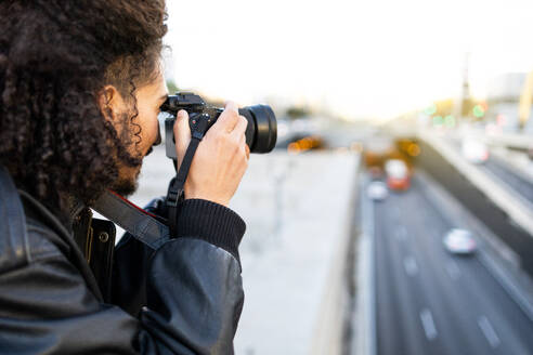 Side view of African American male photographer in trendy leather clothes and curly hair taking picture of motorway with professional photo camera in sunny daylight - ADSF45310