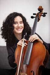 Calm smiling female musician in formal black clothes sitting on sofa and playing acoustic cello with fiddlestick while looking at camera - ADSF45316