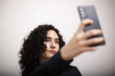 Eine elegante Frau mit lockigem Haar macht ein Selfie mit ihrem Smartphone, während sie in einer formellen schwarzen Jacke auf einem weißen Sofa sitzt - ADSF45318