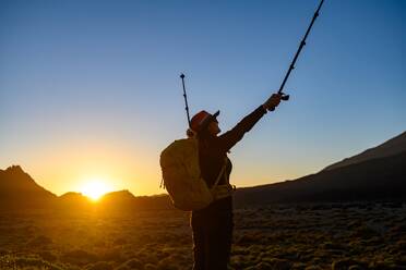 Side view of a woman traveler with a backpack admiring the picturesque sunset, standing with her arms raised against mountains and hills during a trekking trip in Tenerife - ADSF45323