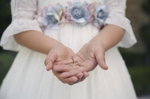 Anonymous girl in white dress with floral decorations demonstrating golden ring during wedding ceremony on blurred background - ADSF45327