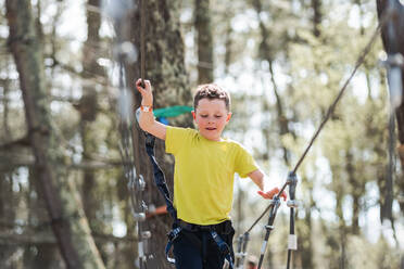 Low angle of positive preteen boy in casual clothes with safety equipment walking on rope bridge against blurred trees in woodland during summer weekend - ADSF45332