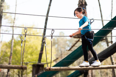 From below full body of a happy and focused preteen girl in casual clothes and safety gear standing balancing while walking on a wooden bridge in the rope park - ADSF45333