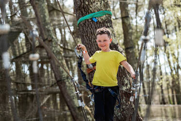 Happy preteen boy in casual clothes and safety gear, walking on a rope bridge against blurry trees in a wood during a summer weekend - ADSF45334