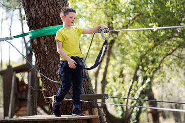 Positive preteen boy in casual clothes and safety gear clinging to the rope of a wooden bridge and looking away as he prepares to cross the bridge in the park during the summer weekend - ADSF45336