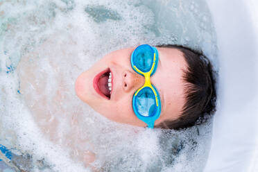 Top view of little boy in blue swimming goggles enjoying bath with bubbles in hot tub in bathroom - ADSF45342