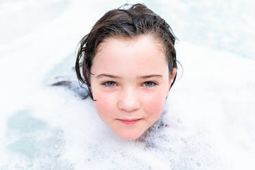 Portrait of cute girl with wet hair looking at camera while relaxing in bathtub with foam during bathing - ADSF45343