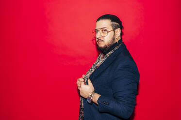 Side view of young bearded dandy guy in expensive outfit with silk scarf and glasses looking at camera while standing against red background - ADSF45358