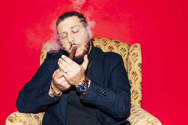 Serious bearded male model in suit smoking cigar while sitting in armchair and blowing smoke against red background in studio - ADSF45366