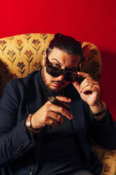 Confident young bearded male model in suit and sunglasses sitting in armchair with cigar and looking at camera against red background in studio - ADSF45368