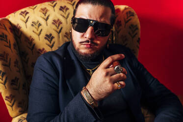 Serious young bearded male model in suit and sunglasses sitting in armchair with cigar and looking at camera against red background - ADSF45369