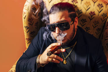 Serious bearded male model in suit smoking cigar while sitting in armchair and blowing smoke against red background in studio - ADSF45370