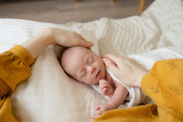 Crop anonymous mother caressing cute newborn sleeping on soft bed in cozy bedroom at home - ADSF45374