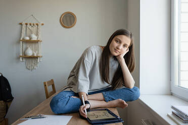 Positive serene teen girl sitting with legs crossed on table and drawing on graphic tablet while looking at camera in own room - ADSF45375