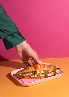 Cropped unrecognizable person eating delicious croissant with ham, cheese and rocket leaves placed on plate in colorful background - ADSF45391