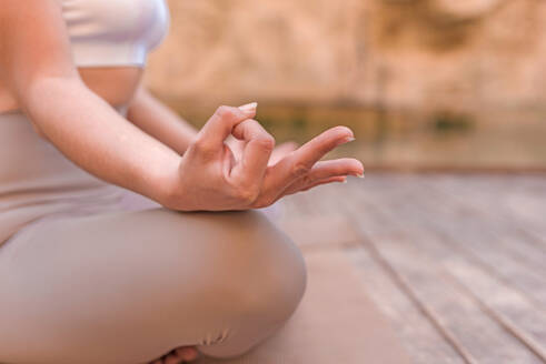 Side view of crop unrecognizable female in sportswear sitting in lotus pose with fingers in mudra while practicing yoga on wooden pier in Charco Azul, Valencia, Spain - ADSF45398