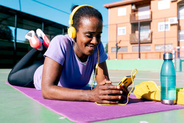 Full length of cheerful black female athlete with dark hair in sportswear smiling and listening to music in yellow headphones headphones via smartphone while lying on mat after outdoor workout - ADSF45428