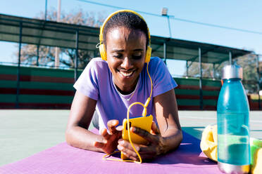 Full length of cheerful black female athlete with dark hair in sportswear smiling and listening to music in yellow headphones headphones via smartphone while lying on mat after outdoor workout - ADSF45429